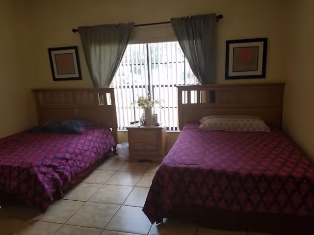 Two twin beds with wooden headboards and matching purple patterned bedspreads flank a nightstand beneath a curtained window in a tiled-floor bedroom.