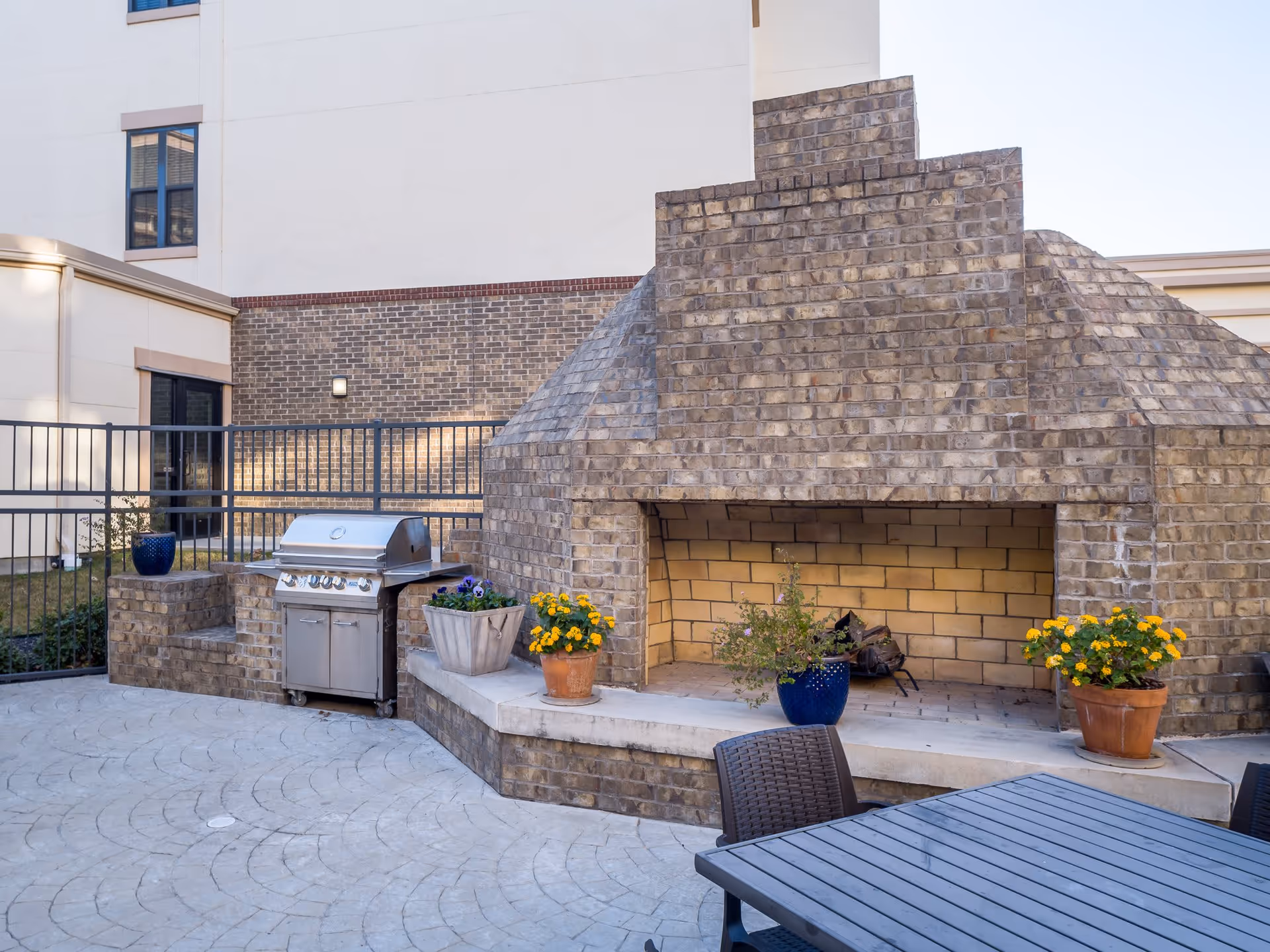 Outdoor patio area with a large brick fireplace, a stainless steel grill, several potted plants with flowers, and a table with chairs. The area is enclosed by a metal fence and adjacent to a building with brick and light-colored walls.