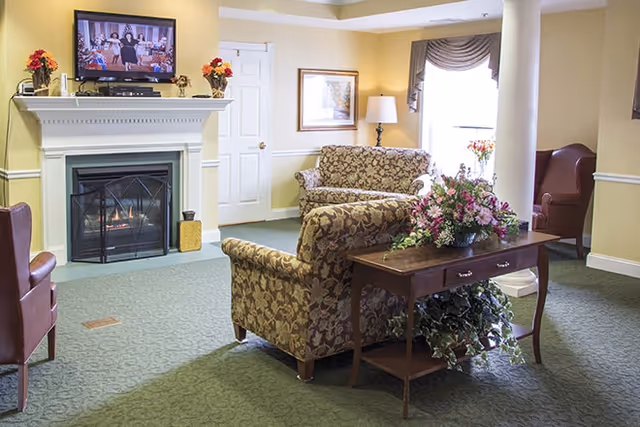 A cozy living room area in a senior living facility with floral patterned sofas, a wooden table with a flower arrangement, a fireplace with a TV mounted above it, and a window with drapes letting in natural light.