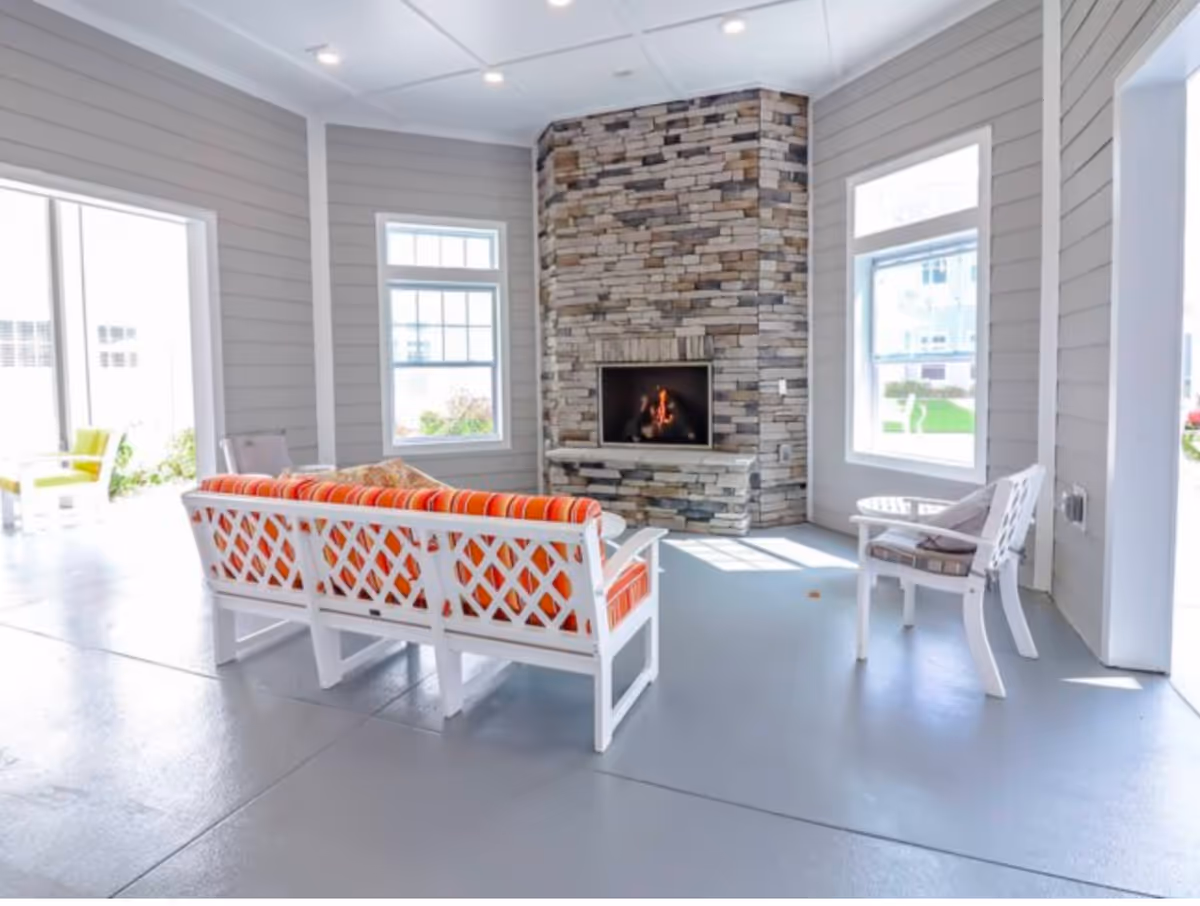 Bright communal living area with white patio-style seating and orange cushions facing a stone fireplace and windows.