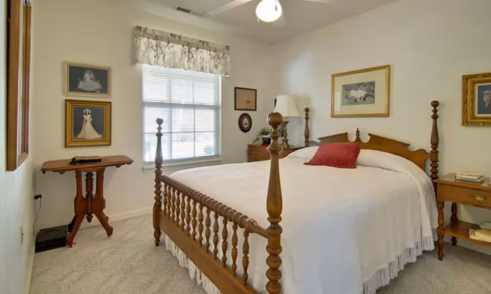 A cozy bedroom with a wooden four-poster bed covered with a white bedspread and a red pillow. The room has beige carpet, a window with blinds and a floral valance, two wooden nightstands with lamps and books, and framed pictures on the walls.
