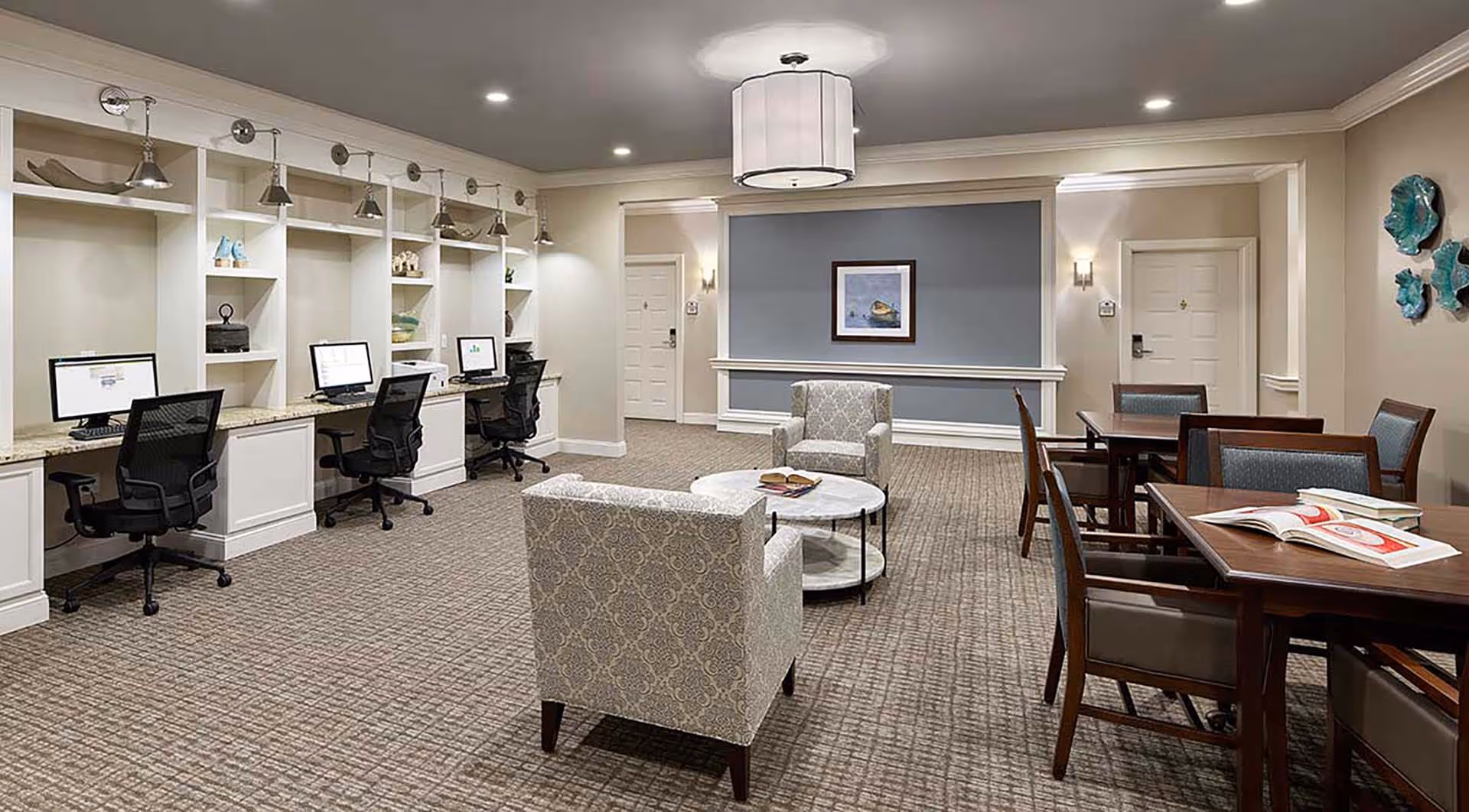 A spacious, well-lit common area in a senior living facility featuring a row of three computer workstations with black mesh chairs along the left wall. In the center, there are two patterned armchairs facing a round marble coffee table with books on it. To the right, there are two wooden tables with chairs, one of which has open books on it. The walls are painted beige with white trim, and a framed picture hangs on a blue accent wall in the background. The ceiling has recessed lighting and a large central light fixture.