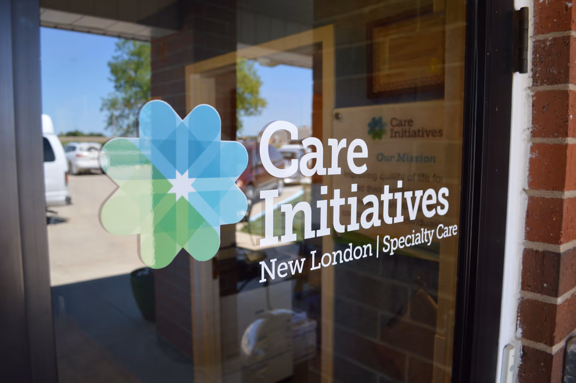 Glass door with the logo and text 'Care Initiatives New London | Specialty Care' printed on it, reflecting the outdoor parking area with cars and trees.