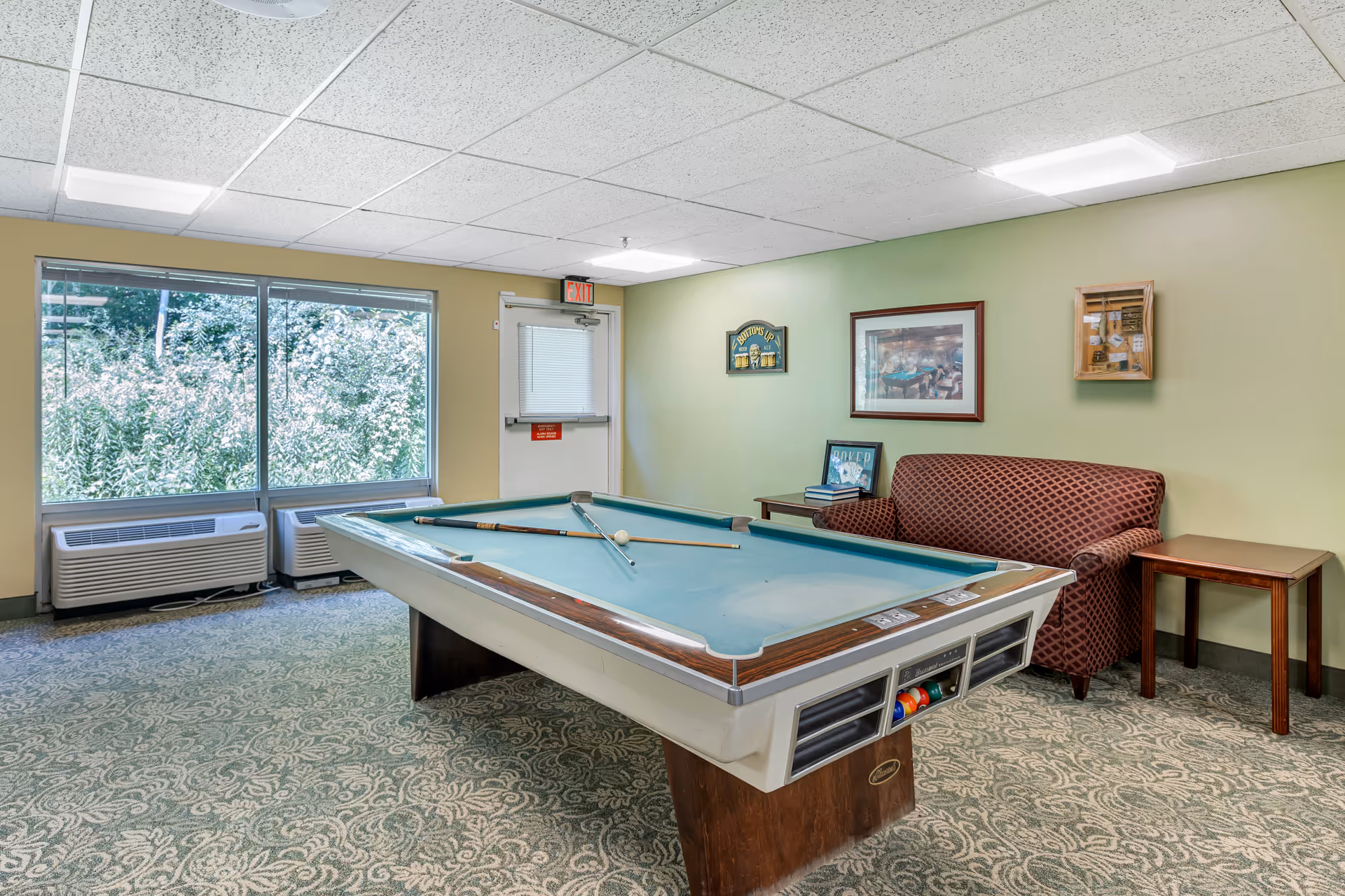 A recreational room with a pool table in the center, two pool cues and a white cue ball on the table. There is a patterned carpet, a maroon patterned loveseat, a small wooden side table, and framed pictures on the light green wall. Large windows show greenery outside, and there is an exit door with a red emergency exit sign above it.