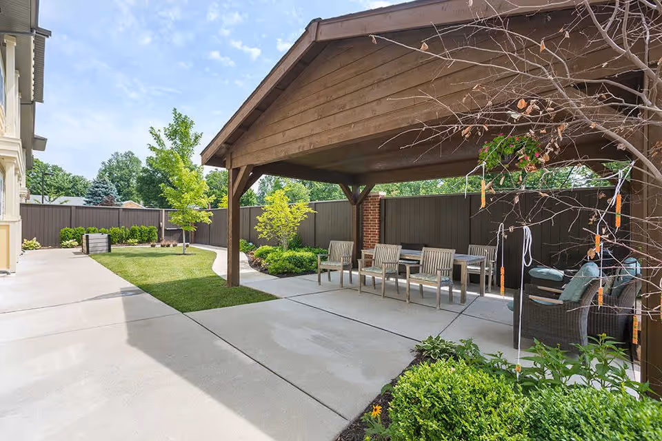 Outdoor patio area at Southview Assisted Living & Memory Care featuring a wooden pergola with seating including chairs and a table, surrounded by a concrete walkway, green lawn, small trees, and shrubs under a partly cloudy sky.