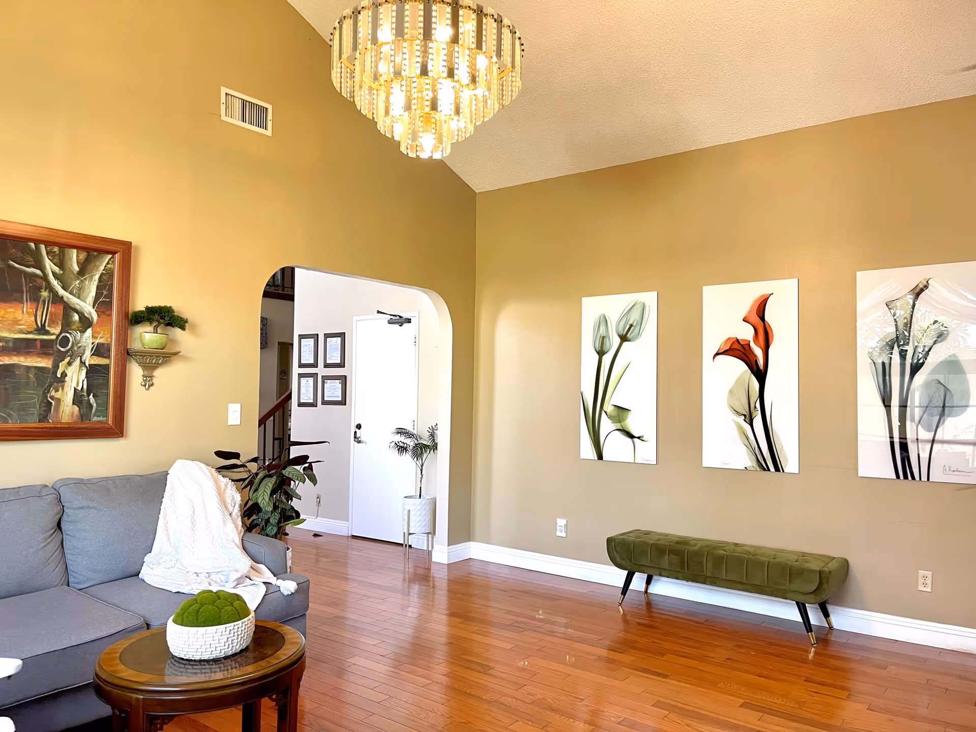 Bright living room with hardwood floors, a gray sofa, green bench, botanical wall art, and a chandelier.