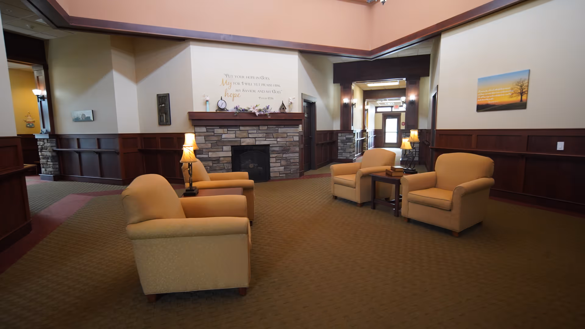 A cozy common area with four beige armchairs arranged around two small wooden side tables with lamps. The room features a stone fireplace with a wooden mantle, decorative items, and a wall decal with an inspirational quote. The walls are painted beige with dark wood paneling, and there is a hallway leading to a door in the background.