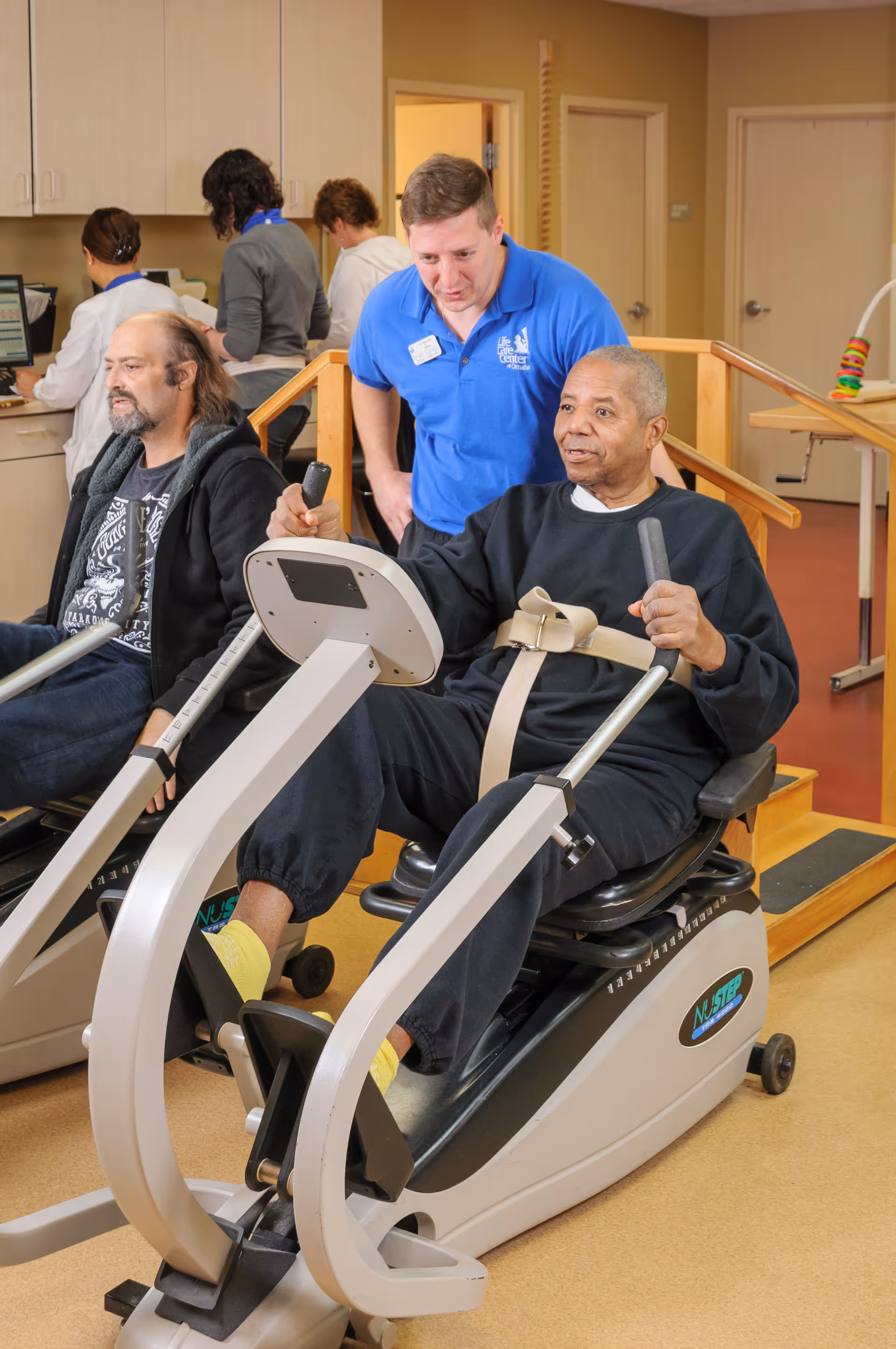 Two elderly men exercising on recumbent stationary bikes in a rehabilitation or senior care facility. A staff member in a blue Life Care Center of Omaha shirt is assisting one of the men. In the background, other staff members are working at a counter with computers.