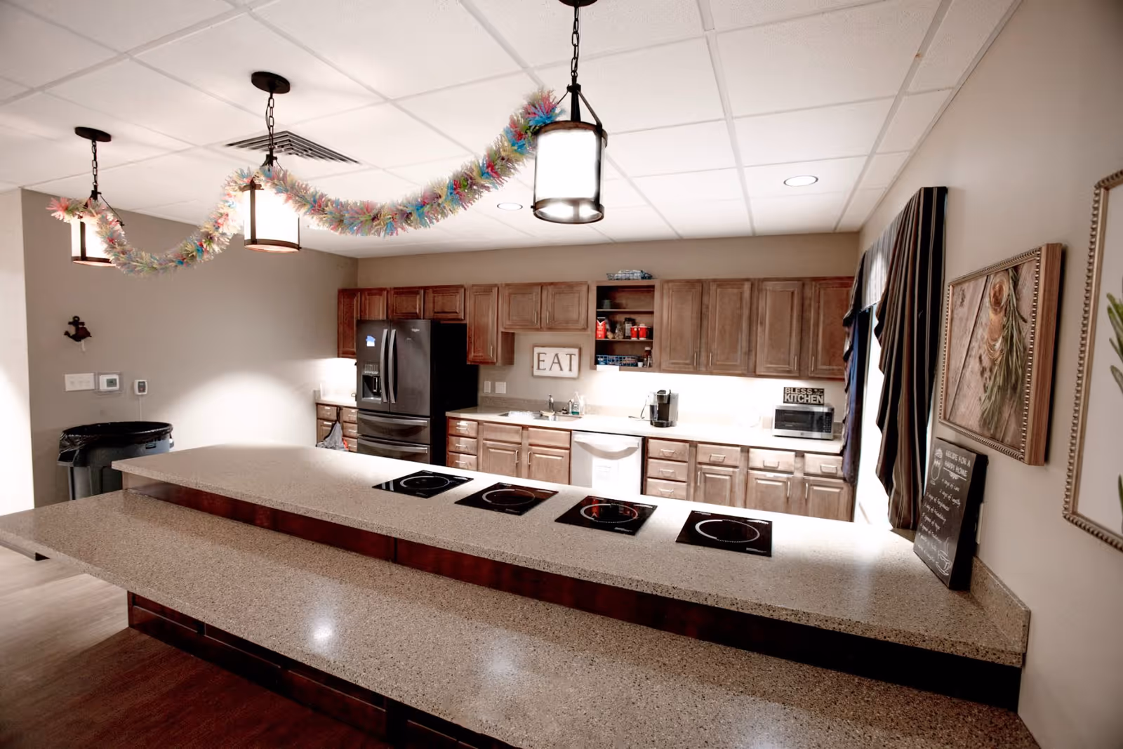 A modern kitchen with wooden cabinets, a black refrigerator, a dishwasher, a microwave, and a countertop with four electric stove burners. The ceiling has three hanging lights decorated with colorful garlands. There are framed pictures and a small chalkboard on the right wall.