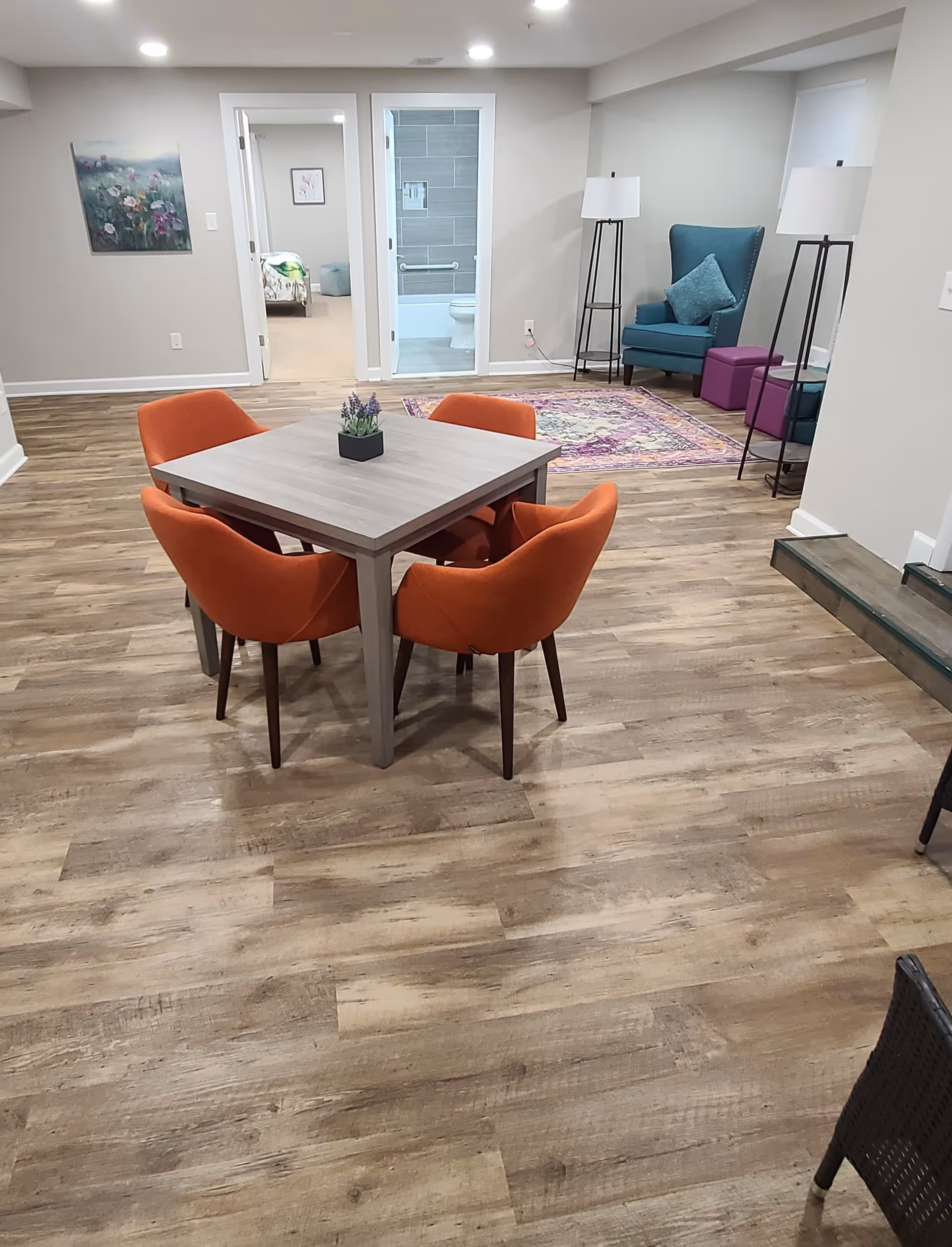 A cozy interior room with a square wooden table surrounded by four orange upholstered chairs. In the background, there is a blue armchair with a matching pillow, two purple ottomans, and two tall floor lamps. A colorful rug is placed near the armchair. There are two doorways visible, one leading to a bedroom and the other to a bathroom with a toilet and grab bars. The floor is wood-patterned, and the walls are painted light gray with a floral painting hanging on one wall.
