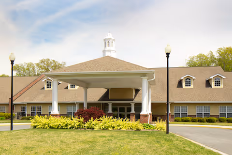 Front entrance of a single-story senior living facility featuring a covered porte-cochère with white columns, a cupola, and a landscaped lawn.