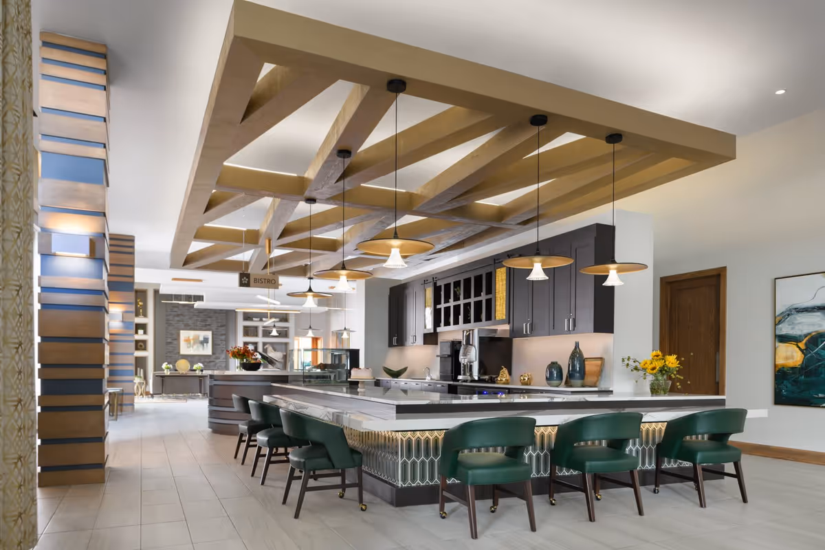 A modern senior living facility bistro area featuring a large marble countertop island with green cushioned chairs on wheels. Above the island, there are multiple pendant lights hanging from a decorative wooden beam ceiling. The back wall has dark cabinetry with various decorative items and kitchen appliances. The space is bright and open with light-colored tiled flooring and contemporary decor.