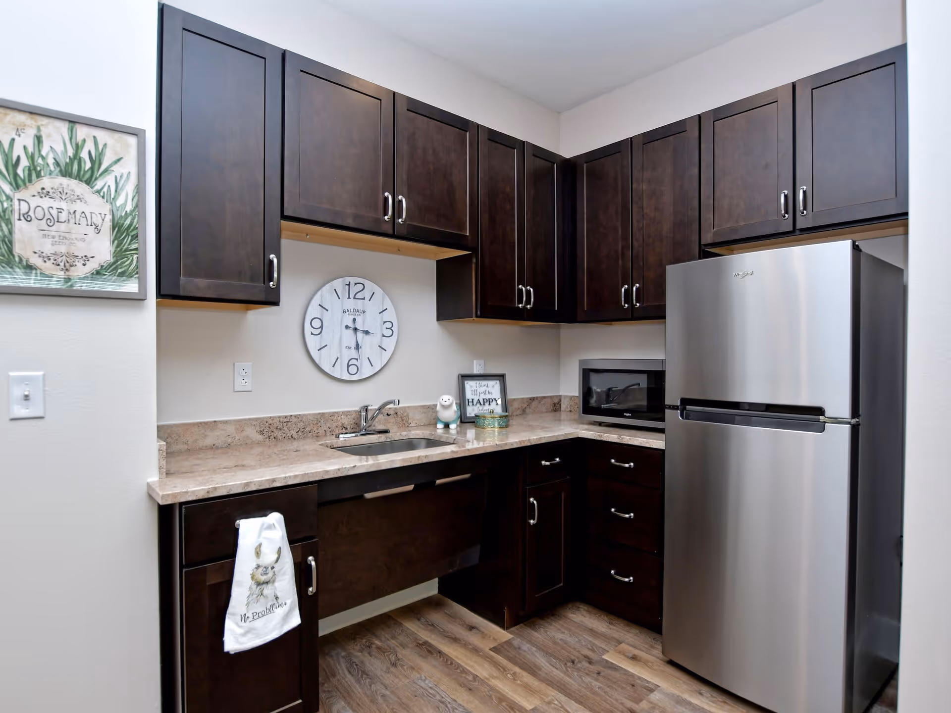 A modern kitchen with dark wood cabinets, a stainless steel refrigerator, a microwave, a sink with a faucet, and a beige countertop. A wall clock and a framed rosemary herb print are hung on the light-colored wall. A decorative towel with a bunny design hangs on one of the cabinet handles.