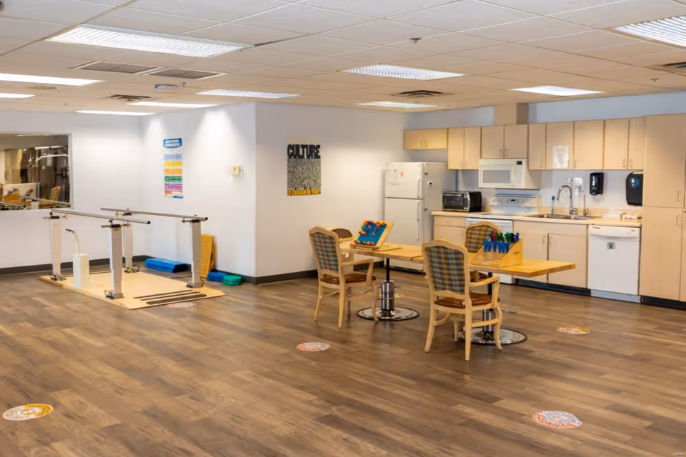 A spacious room with wooden flooring featuring a kitchenette area with cabinets, a refrigerator, microwave, toaster oven, and dishwasher. In the center, there are two tables with chairs and activity games on top. To the left, there is a parallel bar setup for physical therapy or rehabilitation exercises. The walls have posters and the ceiling has fluorescent lighting.