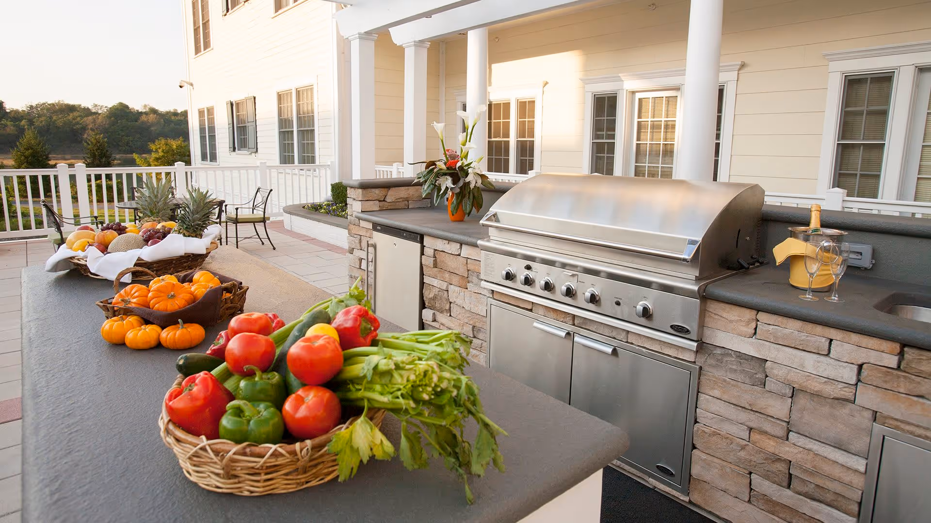 Outdoor kitchen area with a large stainless steel grill, stone countertop, and baskets of fresh vegetables and fruits including tomatoes, bell peppers, celery, pumpkins, and pineapples. There is a bottle of champagne with two glasses on the counter and a flower arrangement near the grill. The area is part of a patio with white railing and chairs, with a view of trees and hills in the background.