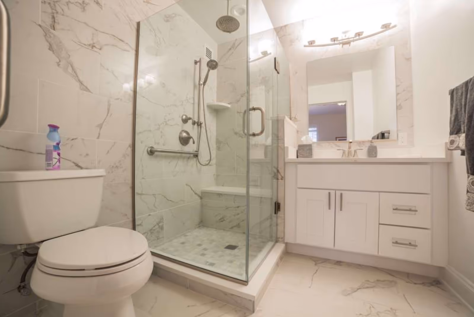 A modern bathroom featuring a glass-enclosed shower with a built-in bench and dual shower heads, a white toilet with a cleaning spray bottle on top, a white vanity with a large mirror and overhead lighting, and marble-patterned tiles on the walls and floor.