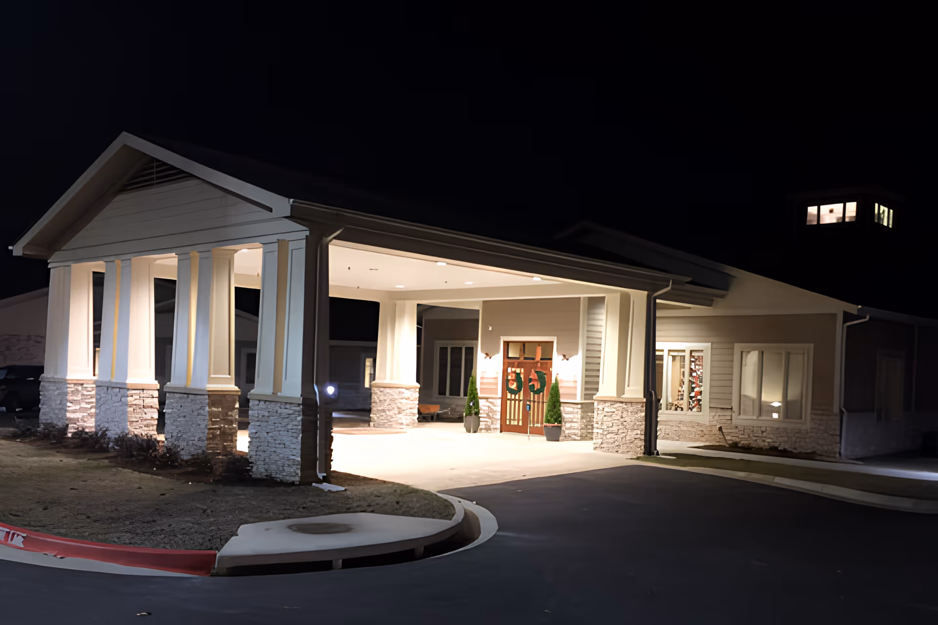 Night view of the entrance to a senior living facility with a covered drop-off area supported by white columns with stone bases. The entrance doors are decorated with holiday wreaths, and the building is illuminated by exterior lights.