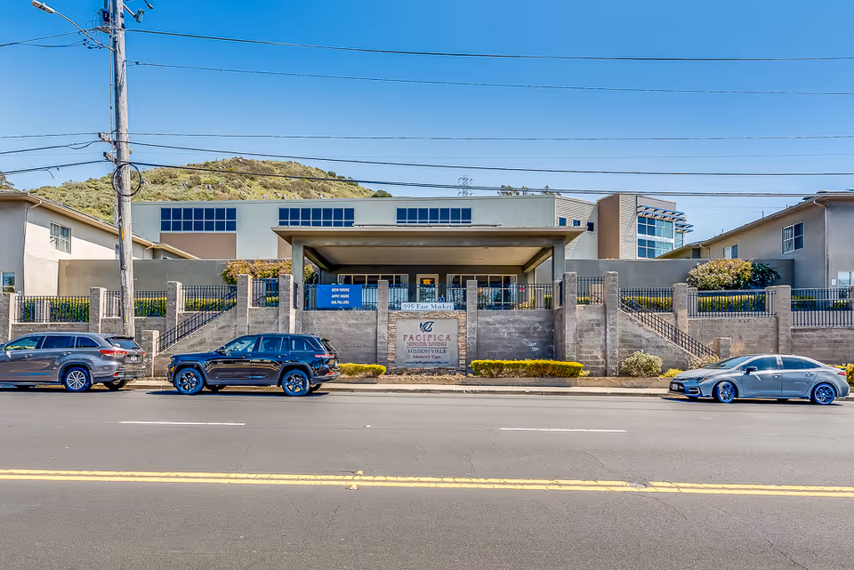 Front exterior view of Mission Villa Senior Living facility with a sign displaying the name and address, three cars parked along the street, and a hill in the background under a clear blue sky.