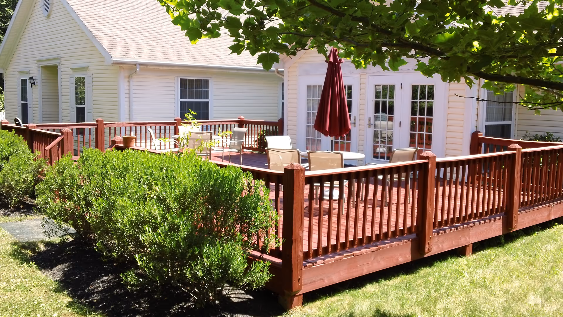 Wooden deck with outdoor dining table and closed red umbrella attached to a light-colored single-story building, with shrubs and grass in the foreground.