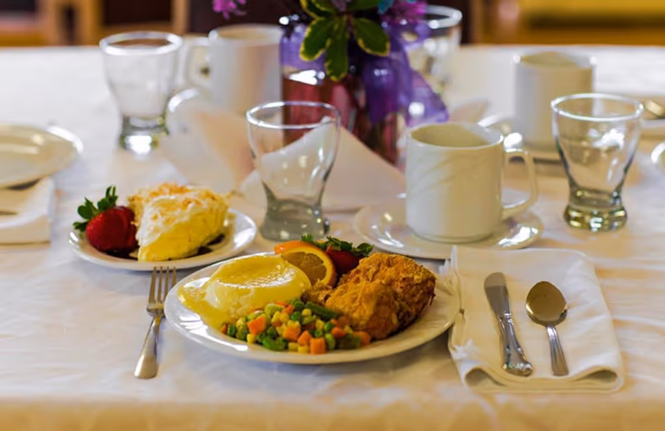 A neatly set dining table with a plate of food including fried chicken, mixed vegetables, mashed potatoes with gravy, and a slice of orange and strawberry. There is also a dessert plate with a slice of pie and a strawberry. The table has white tablecloth, glassware, a white coffee cup, silverware wrapped in a napkin, and a purple flower centerpiece in the background.
