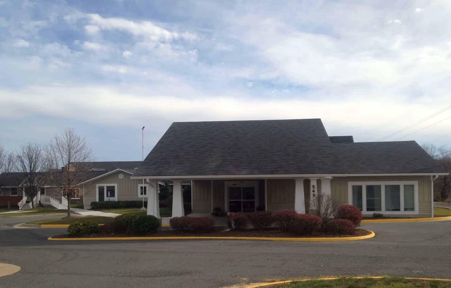 Exterior view of a single-story assisted living facility building with a large covered entrance supported by white columns, surrounded by shrubs and a paved driveway under a partly cloudy sky.