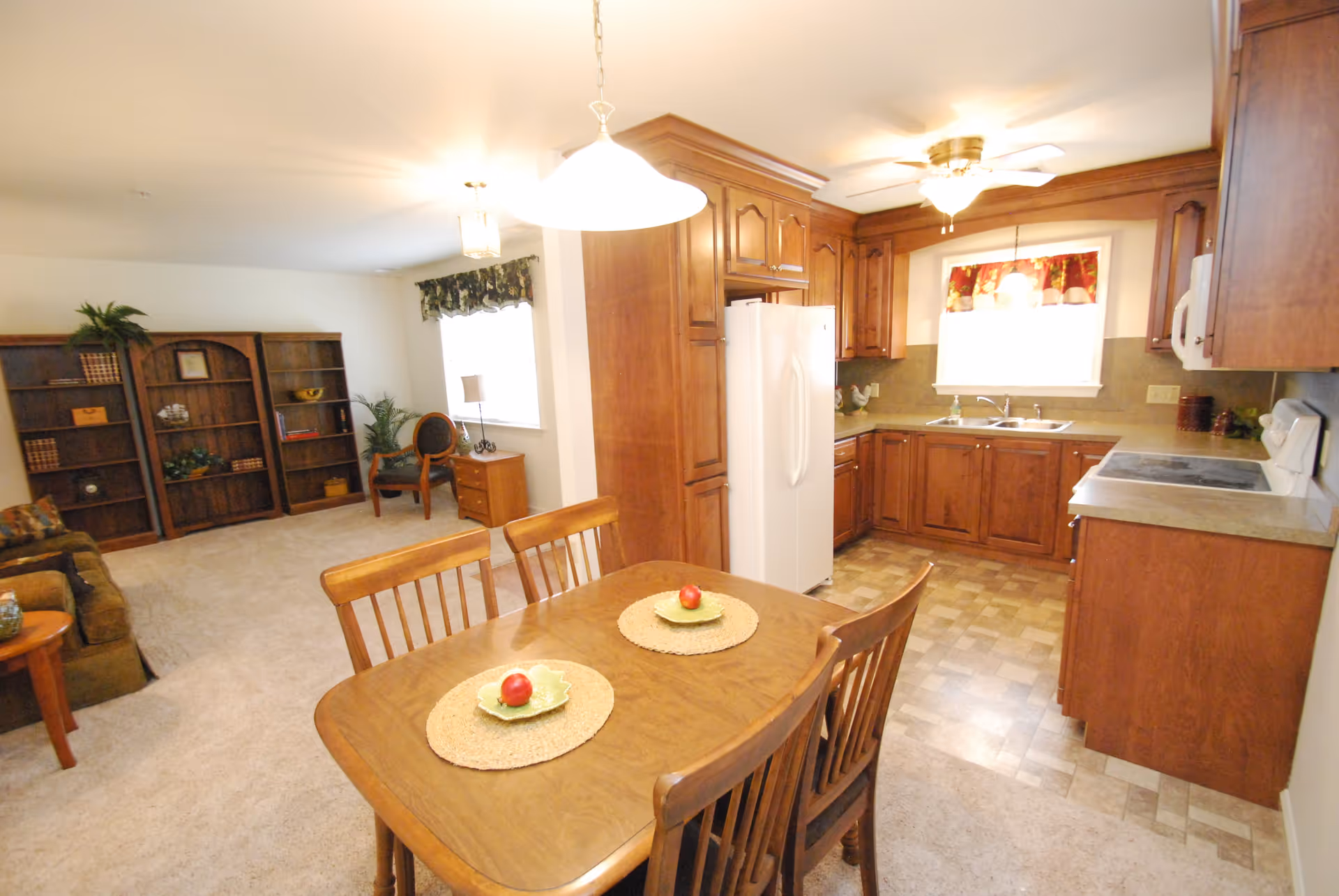 A combined kitchen and living area in a senior living facility. The kitchen features wooden cabinets, a white refrigerator, a stove, and a double sink under a window with a floral valance. A wooden dining table with four chairs is set with two placemats and decorative plates with apples. The living area has carpeted flooring, a brown couch, wooden bookshelves, a chair, and a small side table with a lamp and plant.