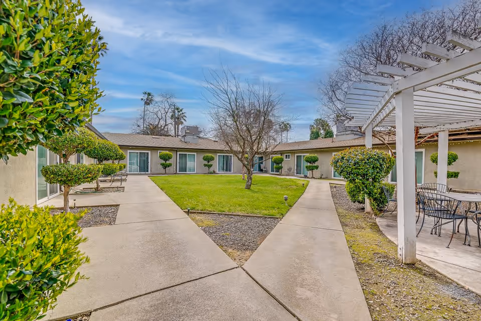 Outdoor courtyard area of Apple Ridge Assisted Living featuring a central grassy lawn surrounded by a paved walkway. The courtyard is bordered by a single-story building with sliding glass doors. There are neatly trimmed bushes and trees, and a white pergola with outdoor seating on the right side.