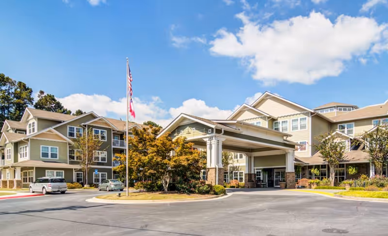 Senior living facility exterior featuring entrance canopy, flagpole, curved driveway, parked cars, and manicured landscaping.