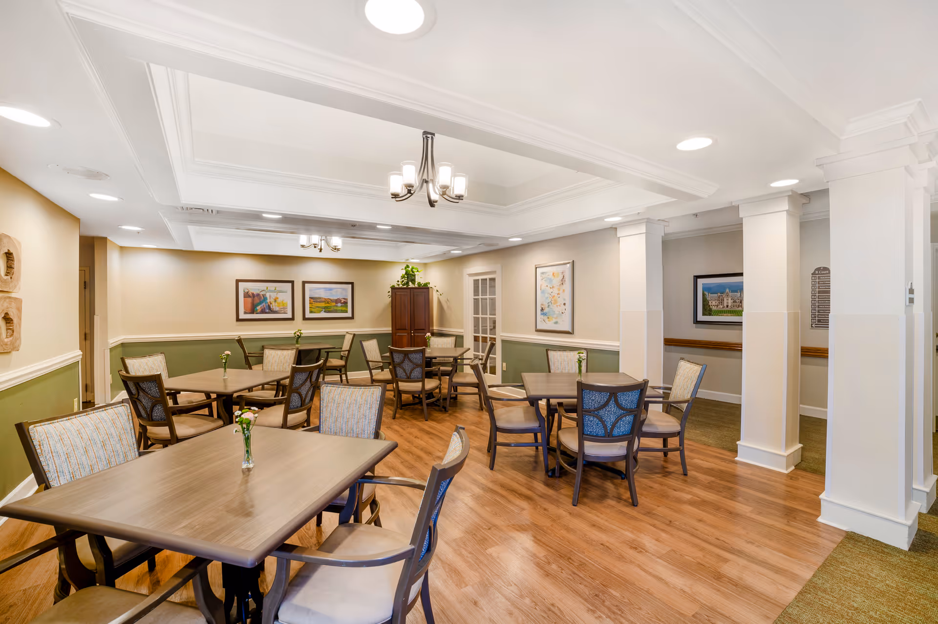 Well-lit communal dining room with several tables and chairs, decorative columns, framed art, and wood flooring.