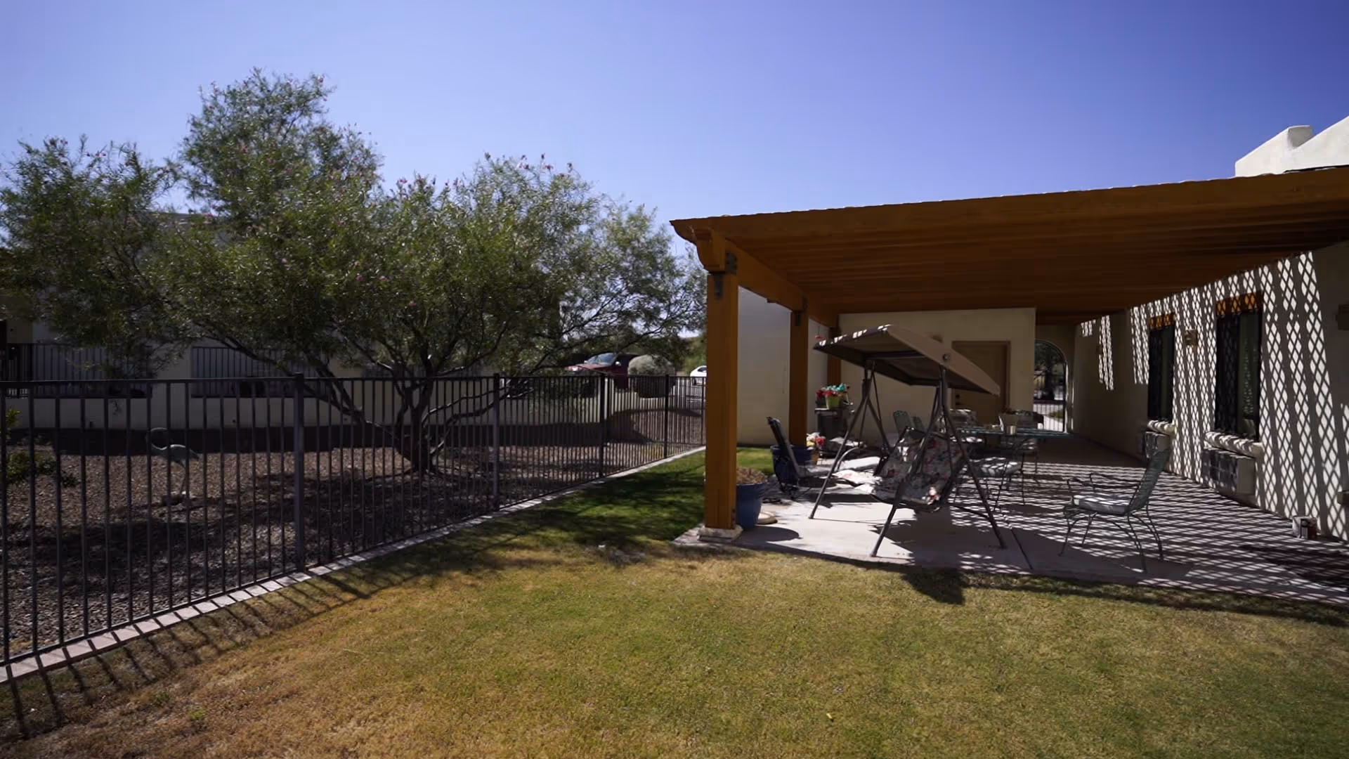 Outdoor patio area at BeeHive Homes of Marana with a wooden pergola providing shade over a swing and several chairs. The patio overlooks a grassy lawn and is adjacent to a fenced area with trees and a clear blue sky above.
