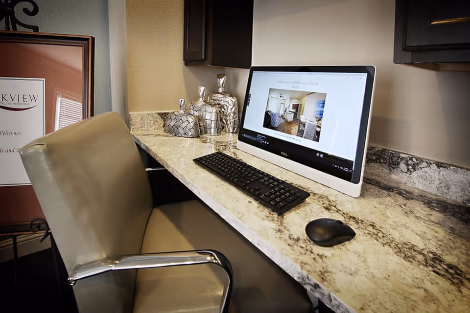 A computer workstation with a Dell all-in-one desktop, keyboard, and mouse on a granite countertop. A beige office chair with metal armrests is positioned in front of the desk. Decorative silver containers are placed on the countertop near the wall. A framed sign for Parkview on Hollybrook is partially visible in the background.
