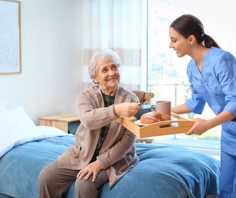 An elderly woman sitting on a bed in a cozy room, smiling and reaching out to take a cup from a caregiver dressed in blue scrubs who is holding a wooden tray with a cup and pastries.