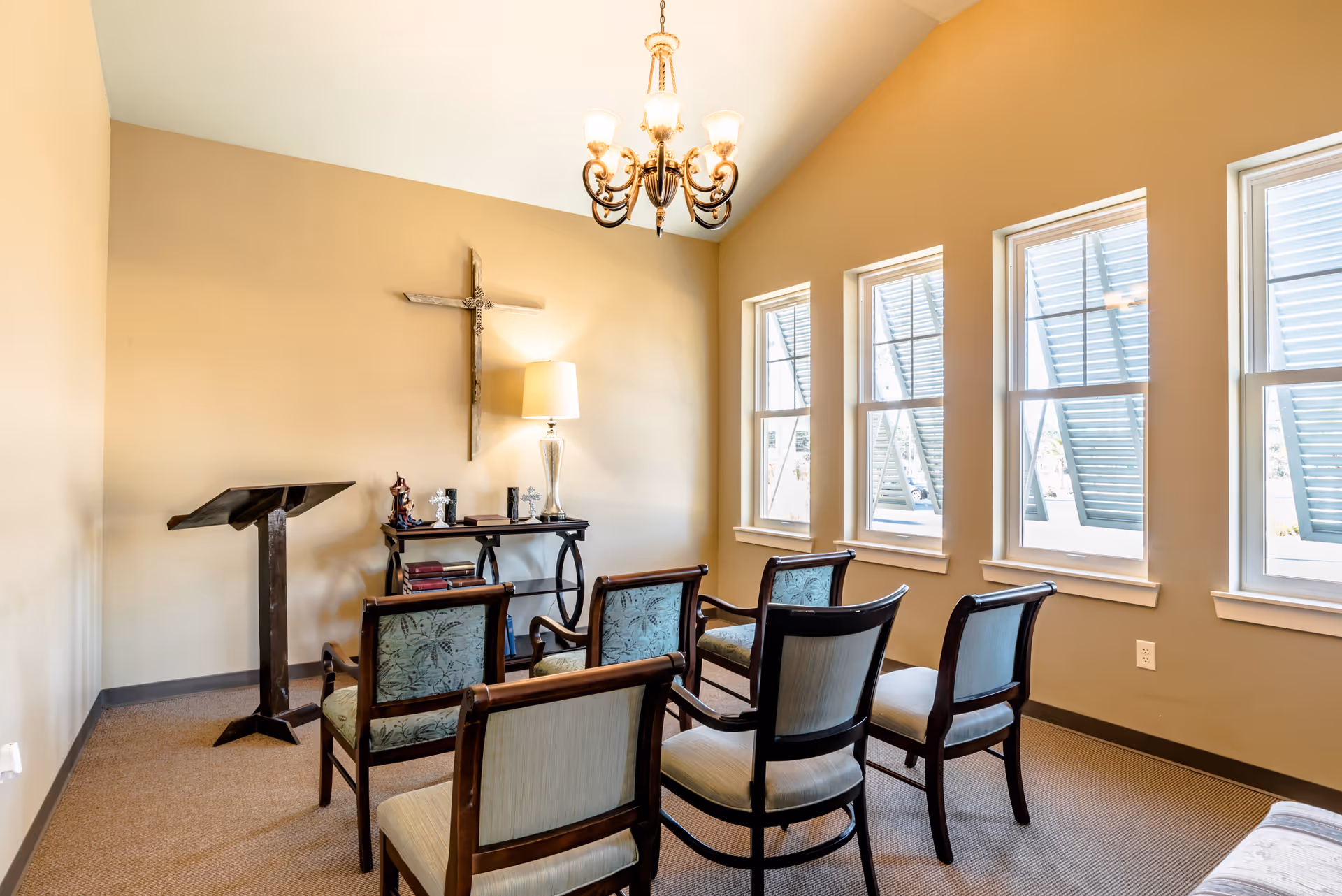 A small chapel or prayer room with six wooden chairs arranged in two rows facing a wooden podium and a table with religious items. A large wooden cross is mounted on the beige wall behind the table, which also holds a lamp and several decorative crosses. Three windows with white shutters allow natural light into the room, and a chandelier hangs from the ceiling.