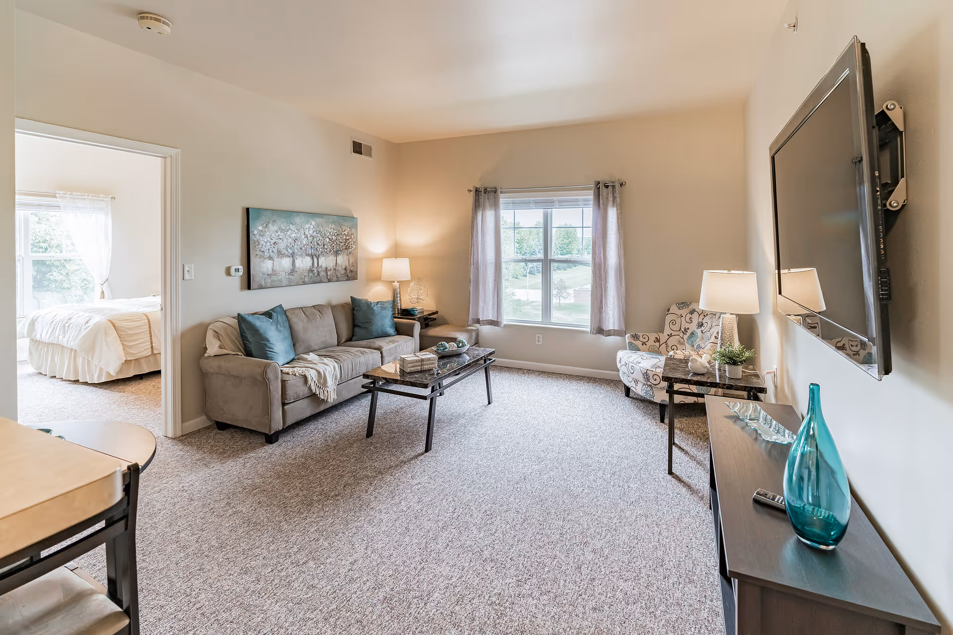 Bright living room with a sofa, coffee table, armchair, wall-mounted TV and a view into a bedroom.