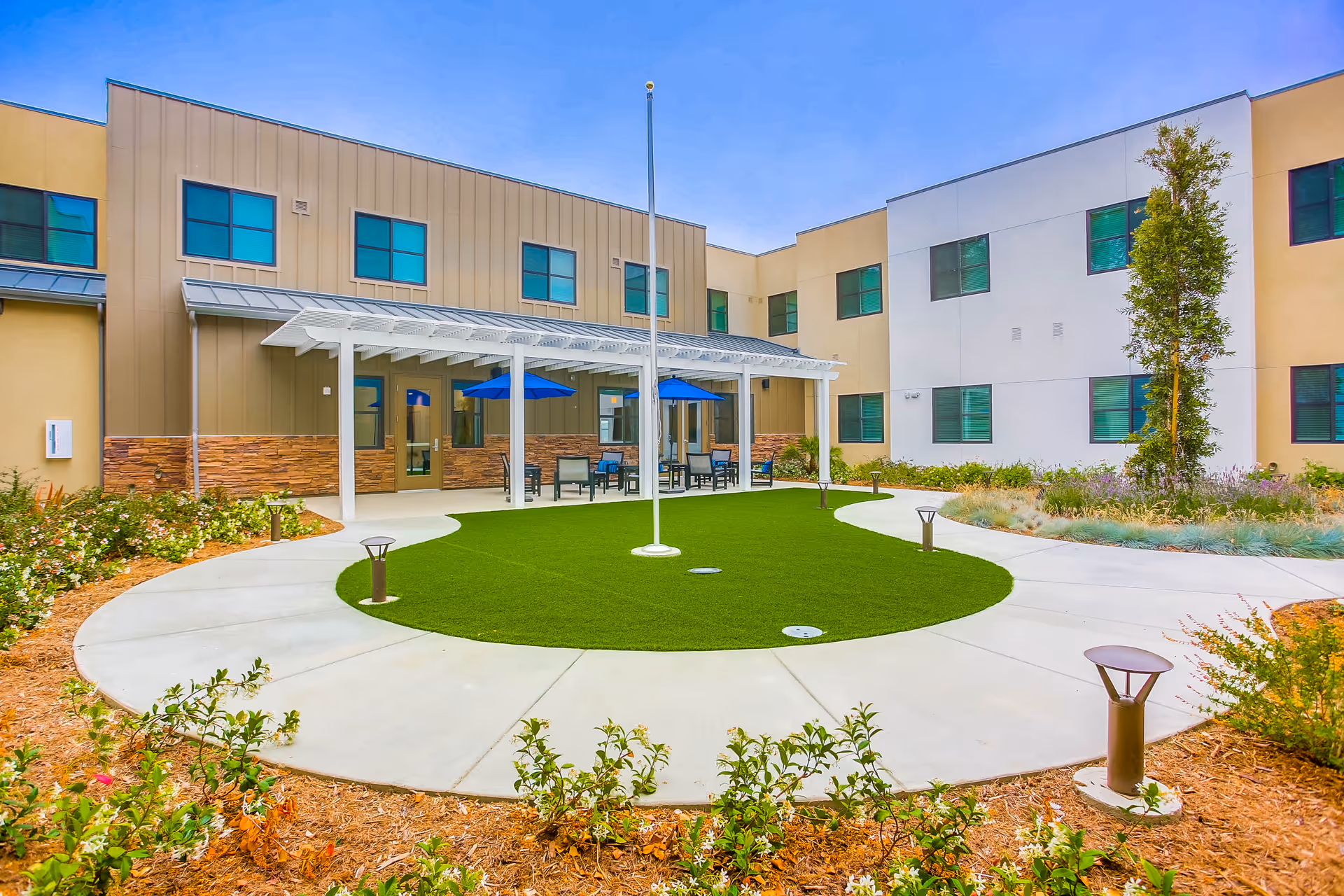 Outdoor courtyard area at Westmont of Encinitas featuring a circular green lawn surrounded by a concrete walkway, garden beds with plants and flowers, a white pergola with blue umbrellas and seating underneath, and a two-story building with multiple windows in the background under a clear blue sky.