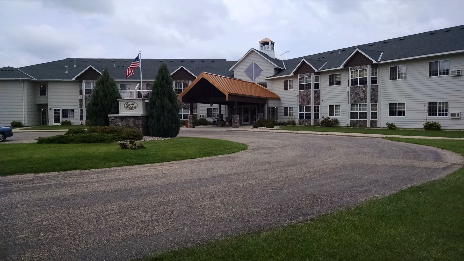 Exterior view of Country View Senior Living by Minnewaska Community Health Services, showing a large two-story building with a covered entrance, an American flag, and well-maintained green lawns and shrubs.