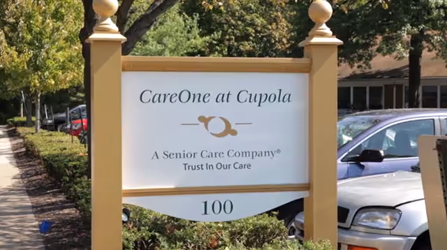 A sign for CareOne at Cupola, a senior care company, with the tagline 'Trust In Our Care' and the number 100 at the bottom. The sign is mounted on two yellow posts with decorative finials, surrounded by greenery and parked cars in the background.