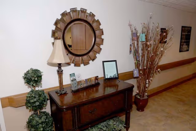 A hallway corner in a senior living facility with a wooden console table against the wall. On the table are a decorative lamp, framed pictures, and a sign that says 'Laugh'. Above the table is a round decorative mirror. To the right of the table is a large vase with tall branches and hanging cards. To the left is a small artificial topiary plant. The hallway has beige walls with wooden chair rails and a tiled floor.