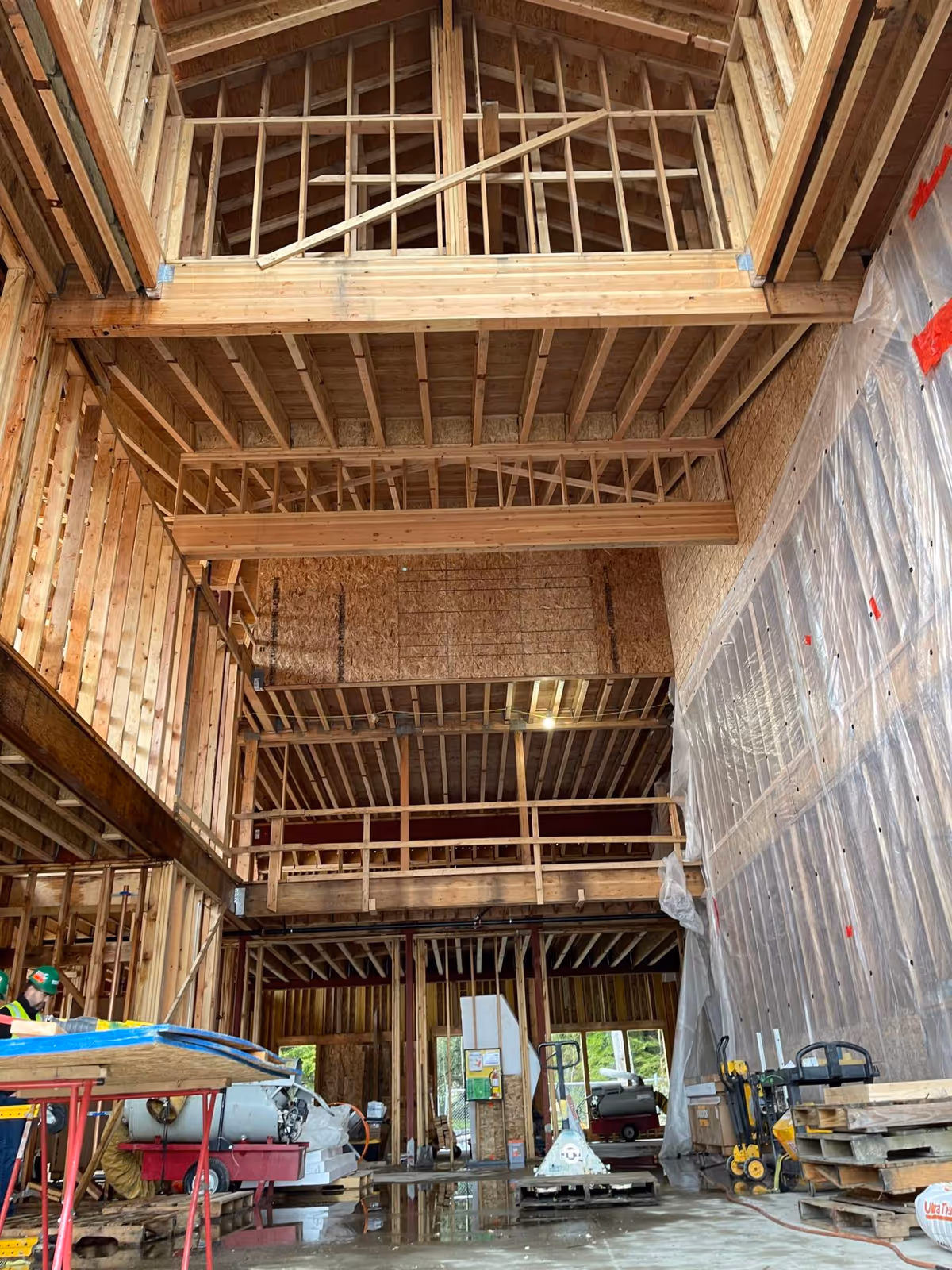 Interior view of a building under construction showing exposed wooden framing and beams, construction materials and equipment scattered on the floor, and a worker wearing a safety vest and helmet on the left side.