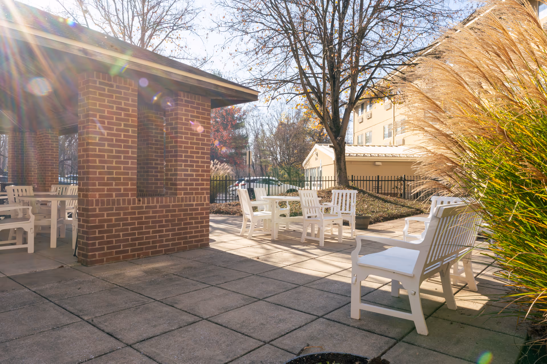 Outdoor patio area with white chairs and tables arranged on a tiled floor next to a brick structure. There are trees with bare branches and ornamental grasses, with a building and a black metal fence in the background under a sunny sky.