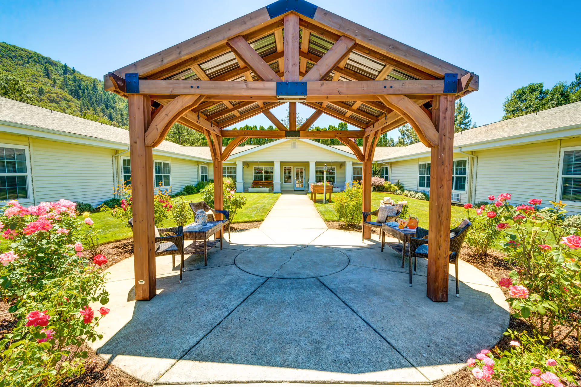 Outdoor seating area with a wooden pergola structure in the center, surrounded by blooming pink and red flowers and green bushes. The area is part of a courtyard with a concrete pathway leading to a single-story building with white siding and multiple windows. The sky is clear and blue, and there are trees and hills in the background.