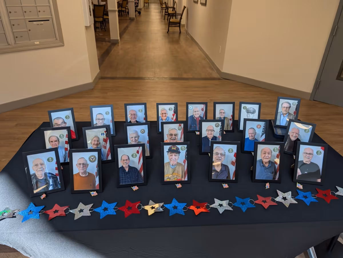 A display table covered with a black cloth featuring framed photographs of elderly individuals, each with a small American flag in the background. In front of the photos, there is a garland made of red, white, and blue star-shaped decorations. The setting appears to be a hallway inside a facility with wooden flooring and beige walls.