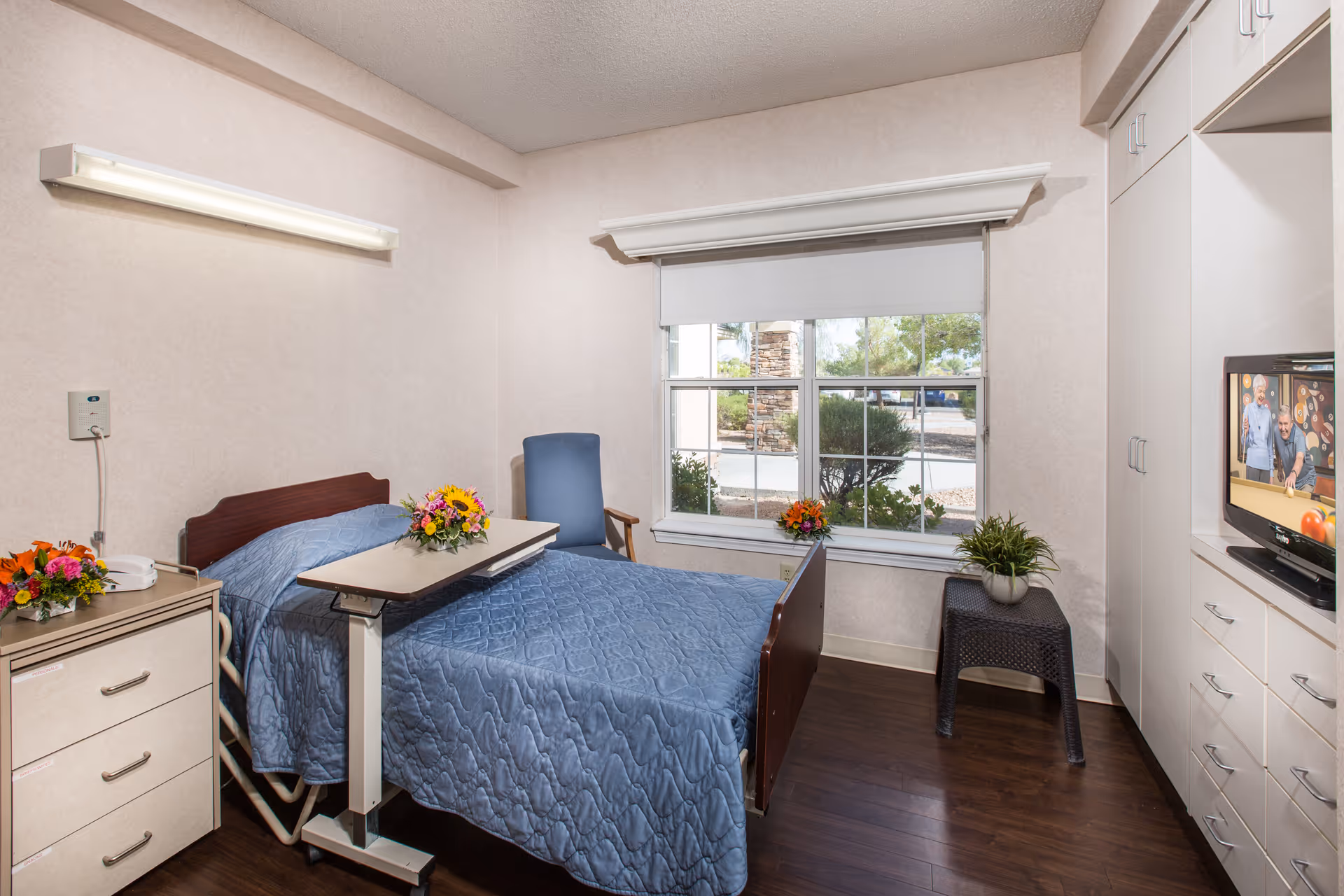 A small, neatly arranged bedroom in a senior living facility featuring a hospital-style bed with a blue quilted bedspread, a bedside table with a floral arrangement and a telephone, a blue armchair, a window with a view of greenery outside, a small table with a potted plant, and a television mounted in a white built-in cabinet showing a billiards game.