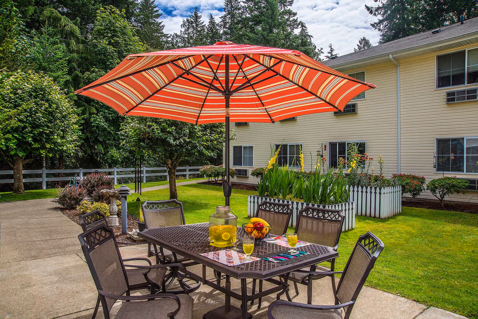 Outdoor patio area with a metal table and six chairs under a large red and orange striped umbrella. On the table are a pitcher of lemonade, three glasses filled with lemonade, and a bowl of fruit. The patio is surrounded by green grass, flower beds with white picket fences, and trees. A beige two-story building with several windows is visible in the background under a partly cloudy sky.