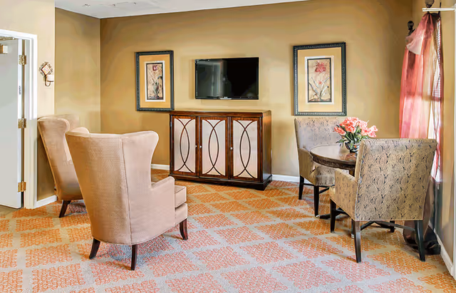 A cozy living room area with two beige armchairs facing a wooden cabinet with a flat-screen TV mounted above it. On either side of the TV are framed floral artworks. To the right, there is a small round table with a flower vase and two patterned chairs near a window with sheer pink curtains.