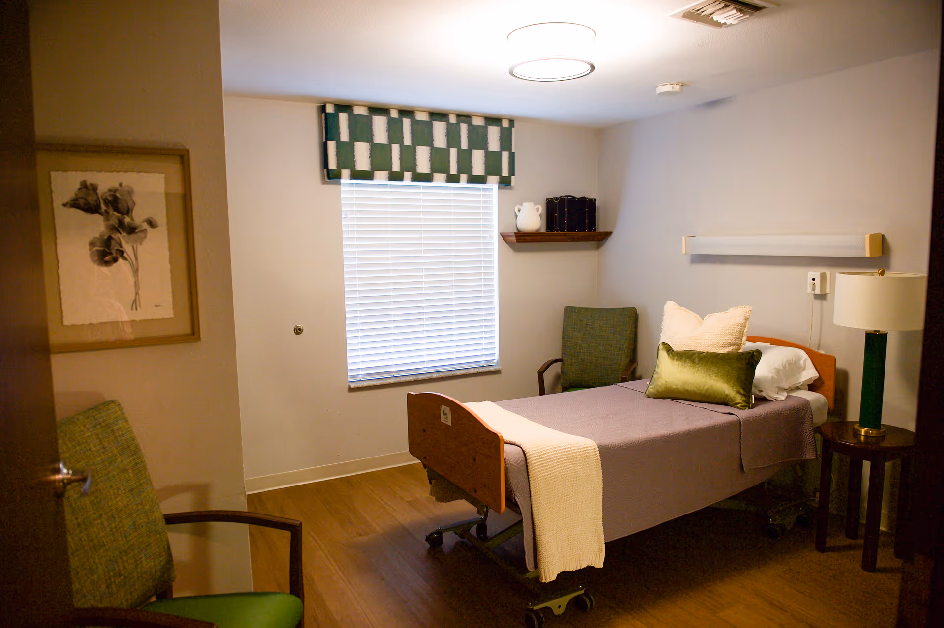 A small, cozy bedroom in a healthcare facility with a single bed covered in a purple blanket and white and green pillows. There is a green armchair near the door, a window with white blinds and a green and white valance, a small wooden side table with a lamp, and a framed floral picture on the wall.