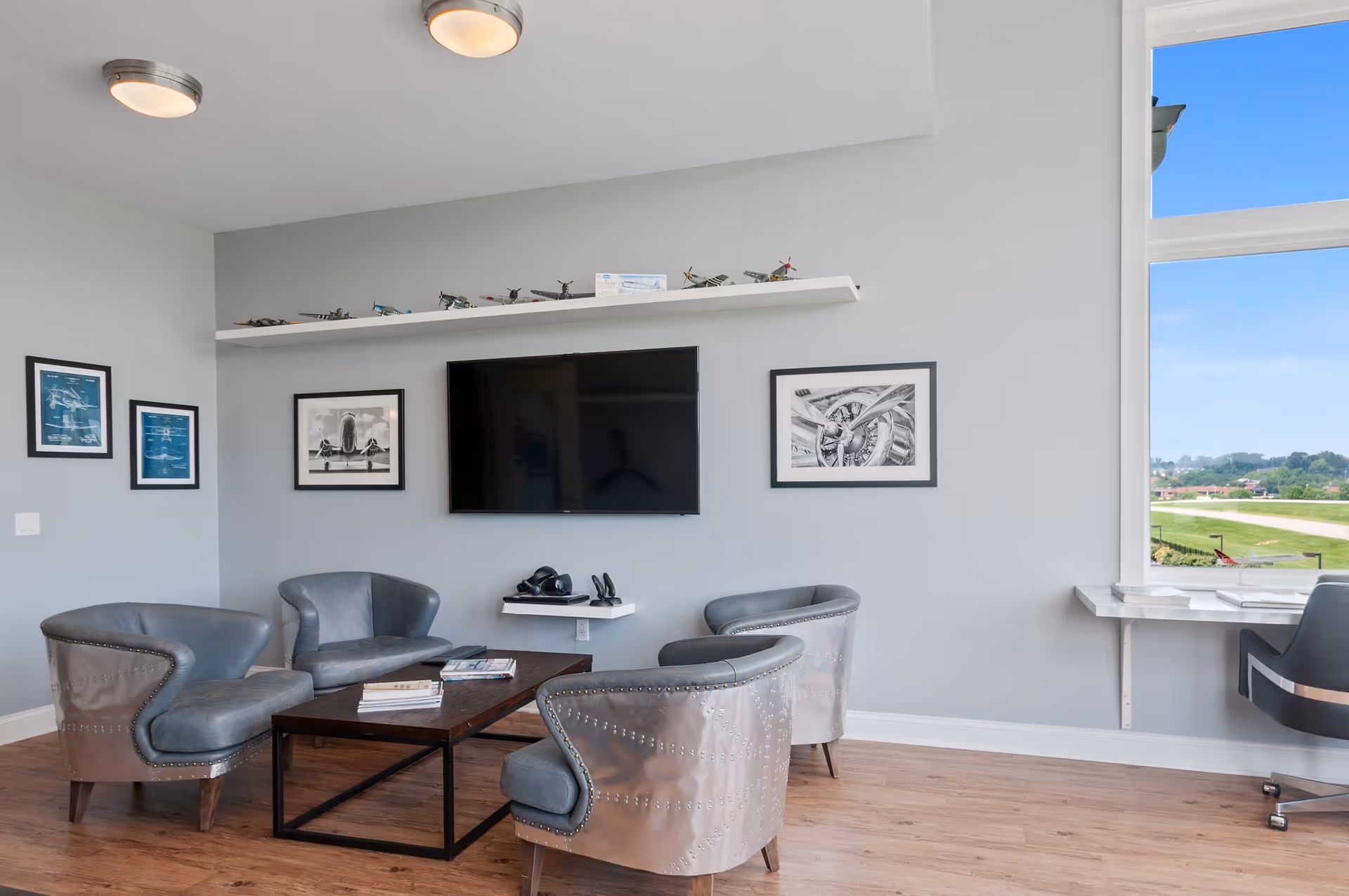A modern lounge area with four metallic armchairs arranged around a rectangular wooden coffee table. A flat-screen TV is mounted on a light gray wall above a small floating shelf holding headphones. Above the TV, a long white shelf displays model airplanes. The walls are decorated with framed black and white aviation-themed pictures. To the right, a large window offers a view of a green landscape and blue sky, with a desk and office chair positioned in front of it. The floor is wooden.