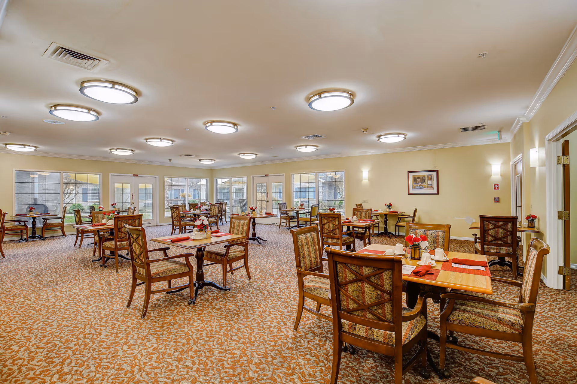 A spacious dining room with multiple wooden tables and upholstered chairs arranged neatly. Each table is set with red napkins, white cups, and small flower arrangements. The room has large windows and glass doors allowing natural light to enter, beige walls, and patterned carpet flooring. Ceiling lights are evenly spaced, providing bright illumination.