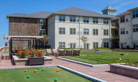 Outdoor courtyard area of Deer Park Village Senior Living featuring a bocce ball court with balls scattered on the green, a well-maintained lawn with chairs arranged around it, flower beds with red flowers, and a multi-story residential building in the background under a clear blue sky.