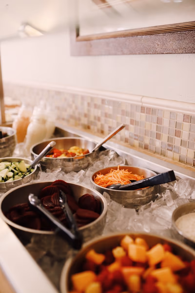 Salad bar with stainless bowls of sliced beets, shredded carrots, cucumbers and fruit set on ice with serving tongs.